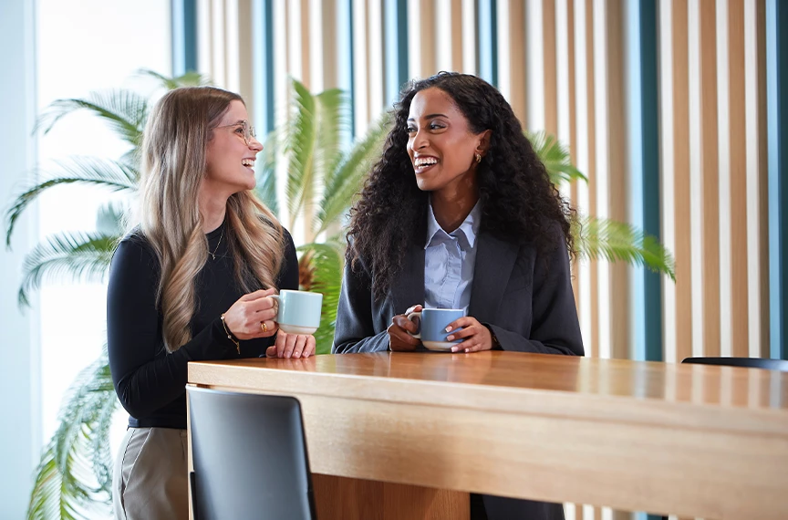 Zwei Frauen stehen entspannt an einem Tisch, halten Tassen und genießen ein gemeinsames Lachen.