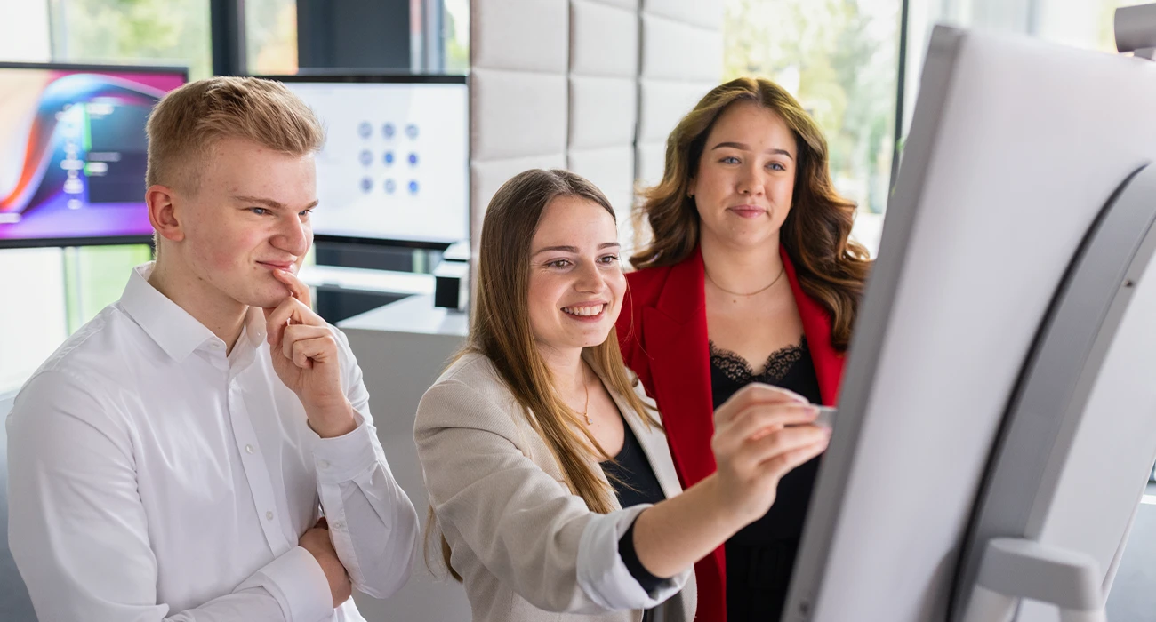The image shows three smiling individuals in front of a flipchart. The woman in the middle is writing something down. 